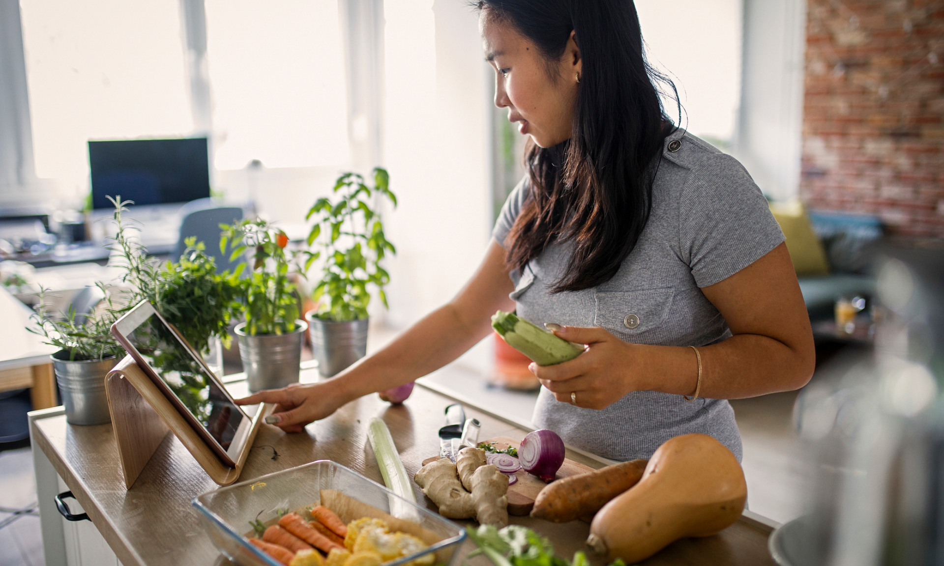 A woman in her kitchen holding a zucchini and referencing a recipe on a tablet