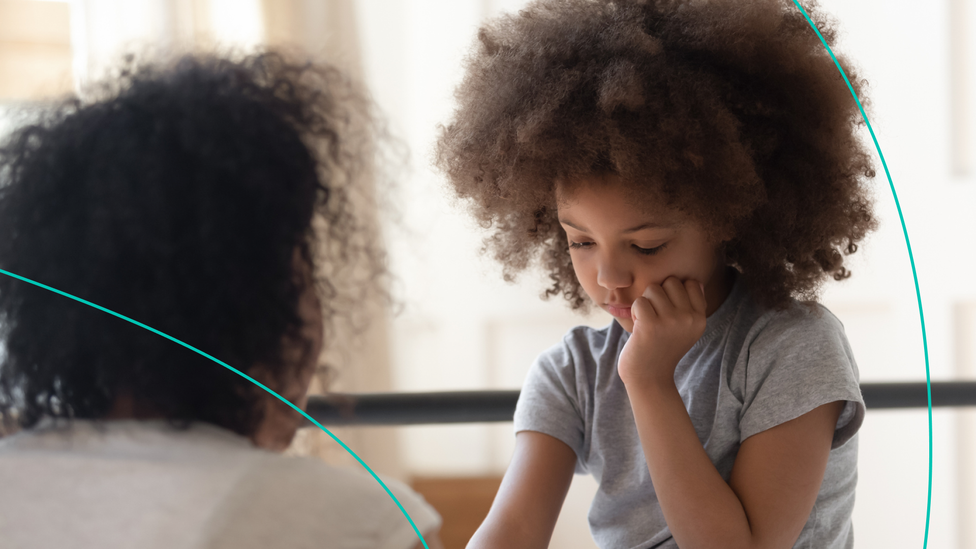 Little girl with curly hair holding her hand to her mouth and looking down sadly while her mom comforts her.